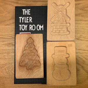 May include: Four wooden cookie cutters, two are rectangular and two are square. The rectangular cutters are a snowman and a windmill. The square cutters are a Christmas tree and a gingerbread house. The cutters are all made of light brown wood.