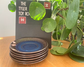 May include: A stack of five blue and brown ceramic saucers with a scalloped edge. The saucers are stacked on a wooden surface.