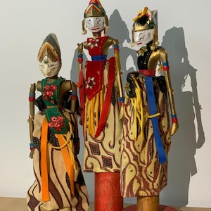 Puede incluir: Tres marionetas de varilla Wayang Golek con rostros blancos, rasgos negros pintados, brazos dorados y ropa de tela. Las marionetas visten faldas de batik estampadas en beige y marrón, acentuadas con cintas rojas, naranjas, azules y amarillas.