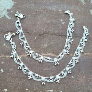 May include: A pair of silver anklets with intricate designs and dangling charms. The anklets are made of multiple layers of delicate chains and have a silver finish.