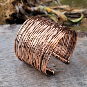 May include: A wide copper cuff bracelet made of multiple thin, hammered bands. The bracelet is open on one side and has a decorative bead at the end.