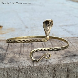 May include: A gold-toned brass bracelet with a cobra snake design. The bracelet is open and has a textured chevron pattern.