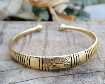 Könnte beinhalten: Ein goldfarbenes Armreif-Armband mit offenem Design. Das Armband hat ein zentrales dekoratives Feld mit geätzten Blattmustern und vertikalen Linien. Die Enden des Armbands haben abgerundete, kugelförmige Spitzen.