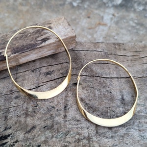 Peut inclure: Une paire de boucles d'oreilles créoles en or avec une forme incurvée légèrement arrondie. Les boucles d'oreilles sont présentées sur une surface en bois.