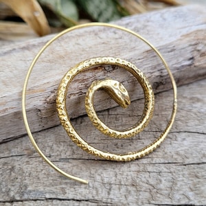 May include: A gold-coloured spiral earring featuring a textured snake head design. The earring is a single piece, coiled into a spiral shape. The snake head is at the centre of the spiral. The earring is set against a wooden background.