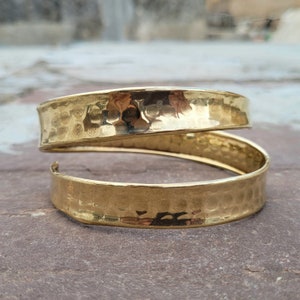 May include: A gold-colored, hammered metal bracelet with a spiral design. The bracelet has a textured surface and a slightly curved shape. The open design allows for easy wear and adjustment. This piece of jewelry is suitable for everyday wear.