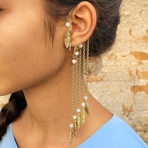 May include: Gold-toned ear cuff featuring a feather design and dangling chains with small white beads. The ear cuff wraps around the ear and has a delicate, bohemian style. The jewelry is shown on a person wearing a light blue top.