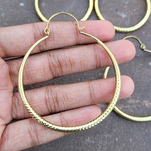 May include: A pair of large, gold-toned hoop earrings with a textured, diamond-shaped pattern. The earrings are shown being held in a hand.