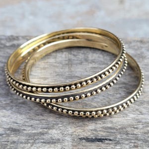 May include: A set of three gold-colored bangles. Each bangle is adorned with a row of small, evenly spaced beads. The bangles are stacked together, creating a layered effect. The jewelry is displayed on a weathered wooden surface.