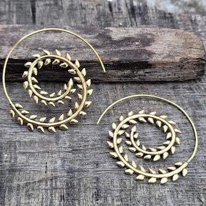 May include: A pair of gold-colored spiral earrings. Each earring features a spiral design with leaf-shaped accents along the outer edge. The earrings are displayed on a wooden surface, showcasing their intricate design and metallic sheen.