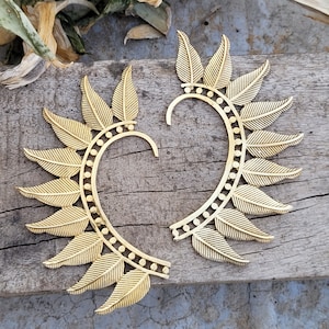 May include: A pair of gold ear cuffs with a leaf design. The cuffs are made of metal and have a delicate, intricate design.