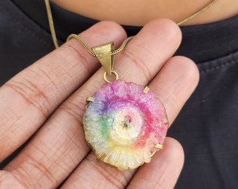 Peut inclure: Un pendentif en agate arc-en-ciel avec un sertissage en laiton. Le pendentif est rond et présente un arc-en-ciel de couleurs au centre. Le pendentif est suspendu à une chaîne dorée.