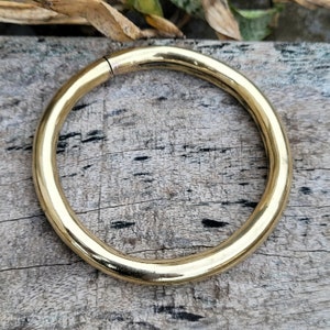 May include: A thick, gold-colored metal ring, likely made of brass, is shown on a wooden surface. The ring is smooth and has a polished finish.