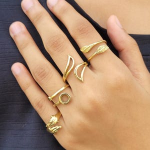 May include: A hand adorned with multiple gold rings. The rings showcase diverse designs, such as leaf patterns, a circular shape, and dolphin figures. The rings are worn on different fingers, highlighting their varied styles and textures.