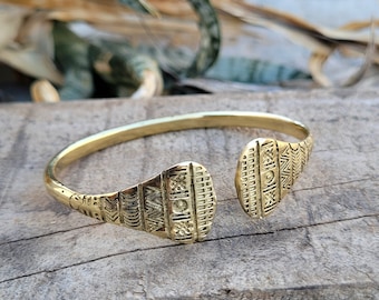 May include: A gold-toned brass bangle bracelet with a geometric pattern. The bracelet is open on one side and has a slightly curved shape.