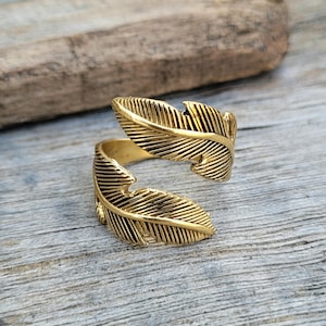 May include: A gold-toned ring with a design of two overlapping feathers. The ring is adjustable and can be worn on any finger.