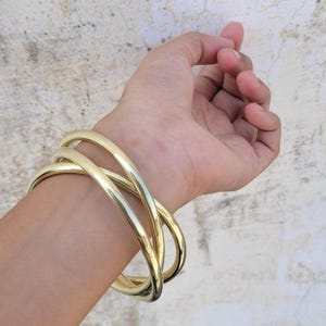May include: A gold-coloured bracelet with a crisscross design. The bracelet is made of thick, rounded metal and is worn on a wrist. The background is a neutral, textured wall. The bracelet is a piece of jewellery.