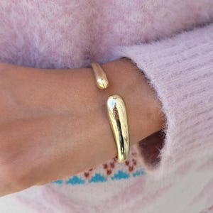 May include: A gold-colored open cuff bracelet. The bracelet has a smooth, polished finish and a teardrop-shaped design at each end. The bracelet is worn on a wrist, against a soft pink sweater.