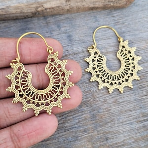 May include: A pair of gold-coloured hoop earrings with an ornate, openwork design. The earrings have a sunburst shape with small decorative elements around the edges. The design includes a curved, open centre with a detailed pattern.