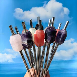 May include: A set of nine reusable drinking straws with different coloured gemstone tops. The straws are made of stainless steel and have a polished finish. The gemstone tops are in shades of pink, purple, and white.