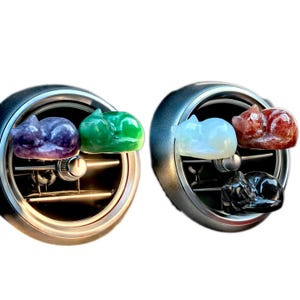 May include: Two car air vent clips with decorative sleeping cat figurines. One clip has a purple and green cat, the other has a white, red, and black cat. The clips are silver and black.