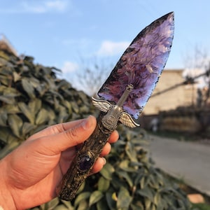 May include: A handcrafted obsidian arrowhead with a purple hue, featuring a decorative handle with a silver guitar and wing design. The handle is adorned with a purple gemstone. The arrowhead is held in a hand.