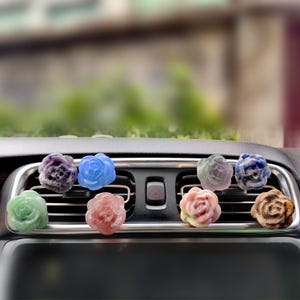 May include: A set of eight car air freshener charms in the shape of roses. The charms are made of different types of stone and are attached to a metal clip. The charms are hanging from the air vent of a car.