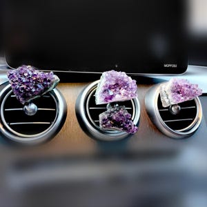 Puede incluir: Tres difusores de ventilación de coche con racimos de cristales de amatista. Los cristales, en varios tonos de púrpura, están montados en rejillas plateadas. El fondo es un interior oscuro y borroso.