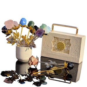 Peut inclure: Un ensemble de sculptures de roses en pierres précieuses dans un vase en céramique blanc et sur une surface réfléchissante. Les roses sont de différentes couleurs, dont le rose, le bleu, le vert et le violet. Une boîte crème avec des détails dorés et le texte "ENJOY THE TIME".