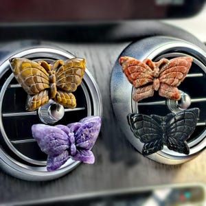 Könnte beinhalten: Vier schmetterlingsförmige Auto-Luftauslassdekorationen. Eine ist bernsteinfarben, eine ist rot-orange, eine ist lila und eine ist schwarz. Jede ist an einer silbernen und schwarzen Autoauslassöffnung befestigt.