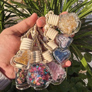 Puede incluir: Una colección de ambientadores decorativos para coche. Los ambientadores tienen recipientes de vidrio transparente en varias formas, incluyendo corazones y una concha. Los recipientes están llenos de piedras y cristales de colores. Cada uno tiene una tapa de madera y un cordón trenzado.