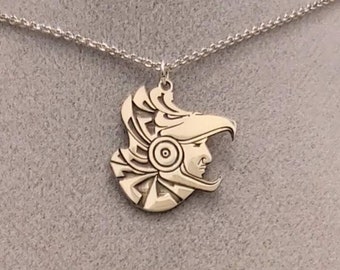 May include: A silver necklace with a pendant featuring a detailed profile of a person wearing a headdress. The pendant is shaped like a crescent moon.