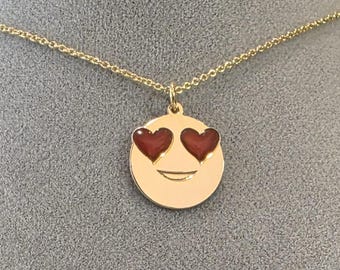 Op de afbeelding: Een goudkleurige ketting met een hanger in de vorm van een emoji-gezicht. De emoji heeft rode hartvormige ogen en een kleine glimlach. De ketting is delicaat en goudkleurig.
