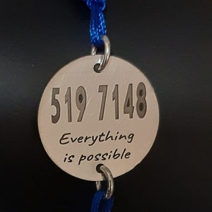 Op de afbeelding: Een blauw gevlochten armband met een zilveren bedel waarop staat "519 7148 Alles is mogelijk".