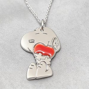 Könnte beinhalten: Eine silberfarbene Metallanhänger-Halskette, die eine stilisierte Zeichentrick-Hundefigur zeigt, die ein leuchtend rotes Herz hält. Der Anhänger hängt an einer zarten silberfarbenen Kette und weist klare Gravurlinien auf, die die Figur definieren.