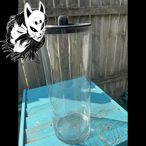 以下が含まれることがあります： 黒いプラスチック製の蓋が付いた透明なガラス製の円筒形のフラワーベース。ベースは青い木製の表面の上に置かれています。