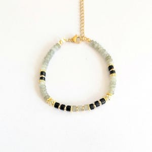 May include: A minimalist bracelet with a gold-coloured chain and clasp. The bracelet features a combination of light green and black beads, with gold-coloured spacers. The beads are small and round, creating a delicate and elegant piece of jewellery. The bracelet is shown against a white background.