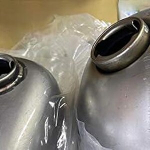 May include: Two silver metal motorcycle gas tanks with round openings for fuel caps. The tanks are unfinished and have a smooth, metallic surface.