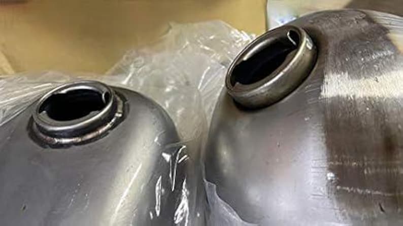 May include: Two silver metal motorcycle gas tanks with round openings for fuel caps. The tanks are unfinished and have a smooth, metallic surface.