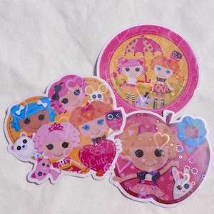 May include: A collection of colorful, die-cut stickers featuring cartoon dolls with large eyes and various hairstyles. The stickers are in the shape of circles and an apple, with the text "Sew Cute" on one.