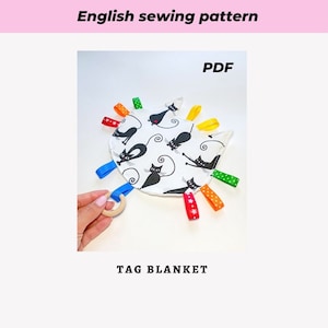 Può includere: Una copertina con etichette bianca con una stampa di gatto nero e etichette di nastro colorate. La copertina ha la forma di una testa di gatto e ha un anello di legno attaccato. Il testo "PDF" e "TAG BLANKET" è stampato sull'immagine.