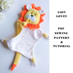 Puede incluir: Un babero de león con una cabeza amarilla y naranja y una manta blanca con puntos dorados. La manta tiene una cola amarilla con una punta naranja. El texto "Lion Lovey" y "PDF Sewing Pattern & Tutorial" está impreso en el lado derecho de la imagen.