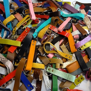Könnte beinhalten: Eine große Sammlung von bunten Leder-Schlüsselanhängern mit Metallringen. Die Schlüsselanhänger sind aus verschiedenen Ledersorten gefertigt, darunter glattes, strukturiertes und geprägtes Leder. Die Schlüsselanhänger haben alle unterschiedliche Längen und Breiten und sind in einem zufälligen Muster angeordnet.