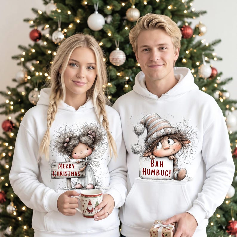 May include: Two white hooded sweatshirts featuring festive illustrations. The left hoodie displays a fairy holding a sign that reads Merry Christmas. The right hoodie shows a character in a striped hat holding a sign that reads Bah Humbug.