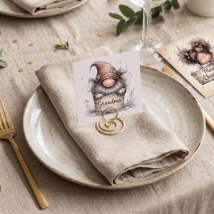 May include: A square place card featuring a gnome illustration with a pink hat and a gray beard, holding a wooden sign with the word "Grandma" printed on it. The card is held upright by a gold-colored spiral metal wire stand on a folded napkin.