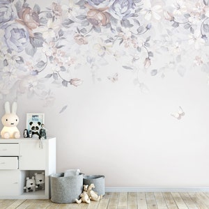May include: A white dresser with two drawers and a white lamp with a bunny rabbit design. There are two grey storage baskets on the floor in front of the dresser. The wall behind the dresser is painted a light purple and features a floral design with butterflies.