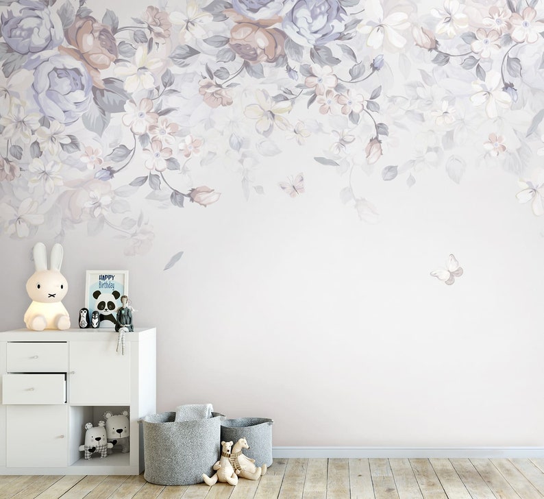 May include: A white dresser with two drawers and a white lamp with a bunny rabbit design. There are two grey storage baskets on the floor in front of the dresser. The wall behind the dresser is painted a light purple and features a floral design with butterflies.