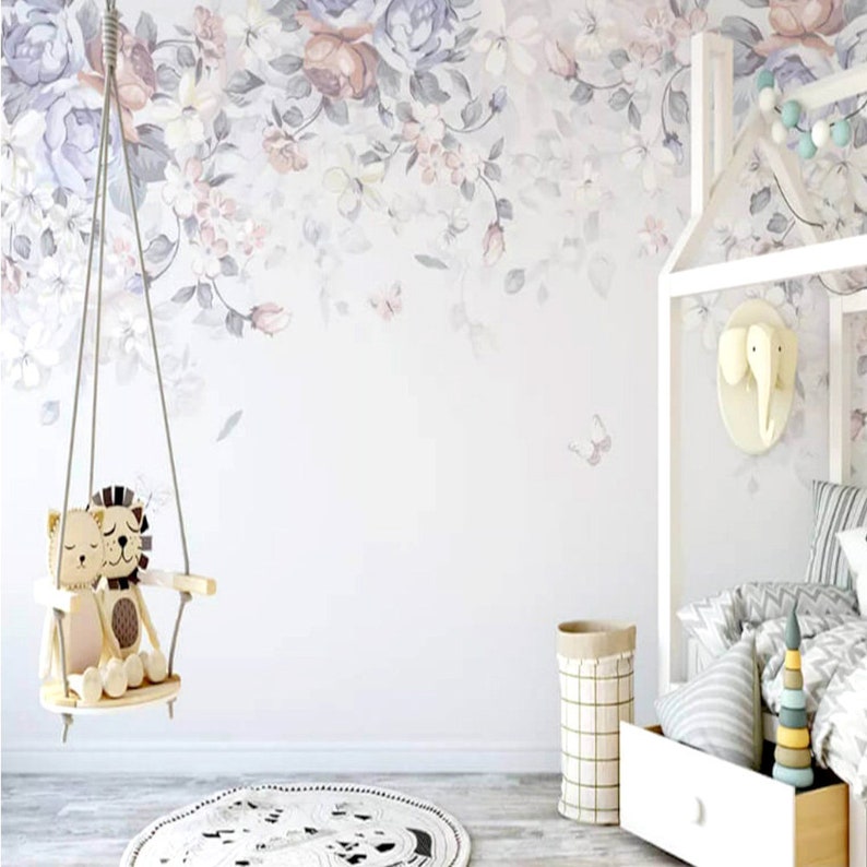 May include: A white wall with a floral wallpaper border. A wooden swing with two stuffed animal lions is hanging from the ceiling. A white cot with a white and grey patterned blanket is in the background. A white wicker basket is on the floor next to the cot.