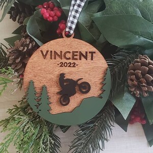 Peut inclure: Une d&eacute;coration de No&euml;l ronde en bois avec un motif vert et marron. La d&eacute;coration pr&eacute;sente une silhouette d'une personne conduisant une moto tout-terrain dans un d&eacute;cor de for&ecirc;t. Le texte "VINCENT -2022- " est grav&eacute; sur la d&eacute;coration.
