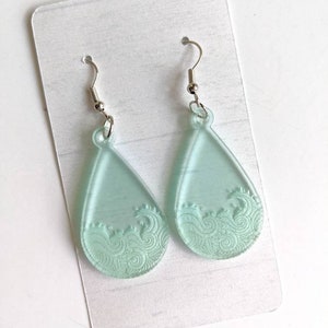 May include: A pair of teardrop-shaped earrings with a frosted, light blue finish. The earrings feature a wave design etched into the front.